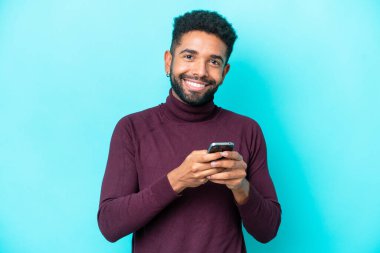 Genç Brezilyalı adam mavi arka planda izole edilmiş cep telefonuyla mesaj gönderiyor.
