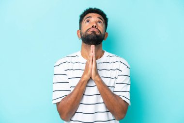 Mavi arka planda izole edilmiş genç Brezilyalı adam avuç içlerini bir arada tutuyor. İnsan bir şey ister.