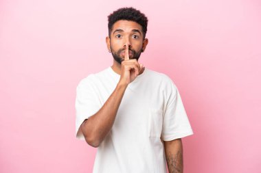 Pembe arka planda izole edilmiş genç Brezilyalı adam sessiz bir jest işareti gösteriyor. Parmağını ağzına sokuyor.