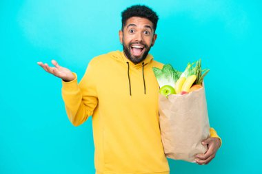 Genç Brezilyalı bir adam mavi arka planda izole edilmiş, şok olmuş bir yüz ifadesiyle paket yiyecek alıyor.