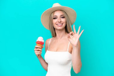Blonde woman in swimsuit holding an ice cream isolated on blue background showing ok sign with fingers
