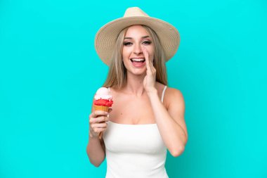 Blonde woman in swimsuit holding an ice cream isolated on blue background shouting with mouth wide open
