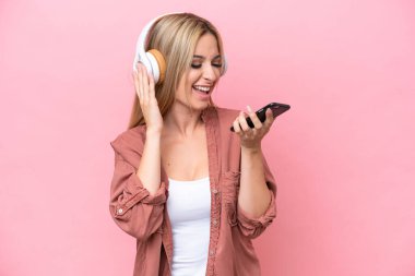 Pretty blonde woman isolated on pink background listening music with a mobile and singing