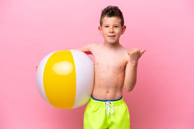 Pembe arka planda izole edilmiş bir plaj topu tutan küçük beyaz çocuk bir ürünü sunmak için yan tarafı işaret ediyor.