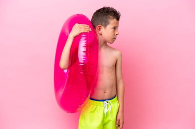 Little caucasian boy holding a inflatable donut isolated on pink background looking to the side