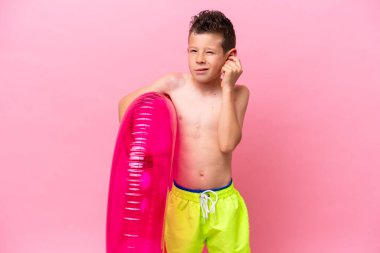 Little caucasian boy holding a inflatable donut isolated on pink background frustrated and covering ears