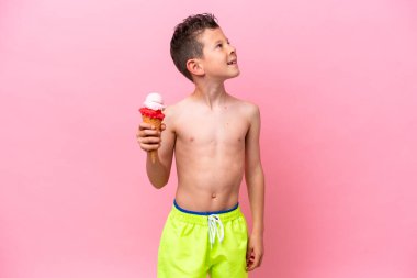 Little caucasian boy eating an ice-cream isolated on pink background looking up while smiling