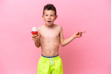 Little caucasian boy eating an ice-cream isolated on pink background surprised and pointing finger to the side