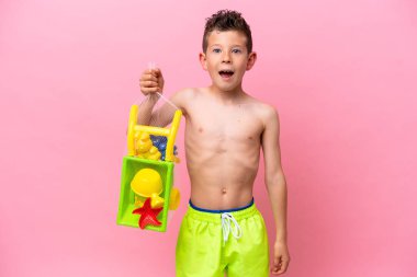 Küçük beyaz çocuk elinde plaj oyuncakları tutuyordu. Pembe arka planda, şaşırmış ve şok olmuş bir yüz ifadesi vardı.