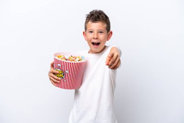 Little caucasian boy isolated on white background holding a big bucket of popcorns while pointing front