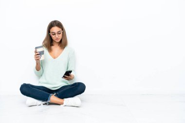 Beyaz arka planda izole bir şekilde yerde oturan beyaz tenli genç bir kadın kahve alıp götürmek için elinde bir cep telefonu...