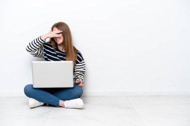 Young caucasian woman with a laptop sitting on the floor isolated on white background covering eyes by hands and smiling
