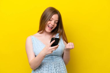 Sarı arka planda izole edilmiş, telefon zafer pozisyonunda genç beyaz kadın.