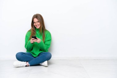Beyaz arka planda izole bir şekilde yerde oturan beyaz bir kadın cep telefonuyla bir mesaj gönderiyor.