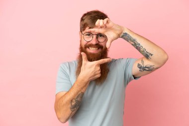 Young reddish caucasian man isolated on pink background focusing face. Framing symbol