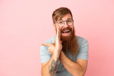 Young reddish caucasian man isolated on pink background pointing to the side to present a product and whispering something