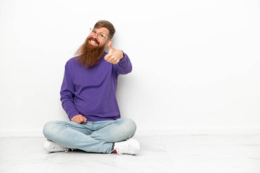 Beyaz arka planda izole bir şekilde yerde oturan Kafkasyalı genç adam. Başparmakları havada. Çünkü iyi bir şey oldu.