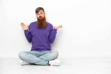 Beyaz arka planda izole bir şekilde yerde oturan beyaz tenli genç adam ellerini kaldırırken şüphe duyuyor.