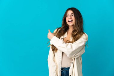 Mavi arka planda izole edilmiş genç beyaz kadın şaşırmış ve işaret ediyor.