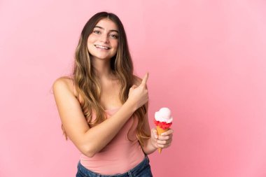 Kornet dondurması olan beyaz bir kadın pembe arka planda izole edilmiş bir şekilde bir ürün sunmak için kenarı işaret ediyor.