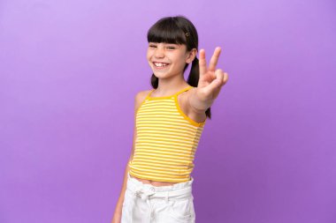 Mor arka planda izole edilmiş küçük beyaz çocuk gülümsüyor ve zafer işareti gösteriyor.