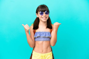 Küçük beyaz bir çocuk kumsala gidiyor. Mavi arka planda izole bir şekilde. Baş parmakları havada ve gülümsüyor.