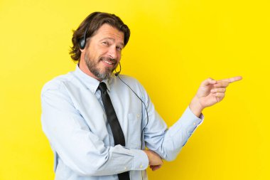 Telemarketer dutch man working with a headset isolated on yellow background pointing finger to the side