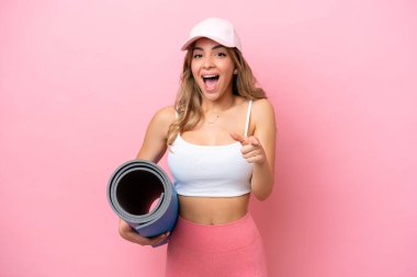 Young sport woman going to yoga classes while holding a mat surprised and pointing front