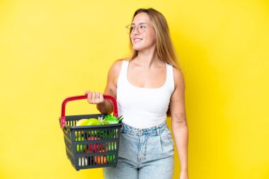 Sarı arka planda izole edilmiş yiyecek dolu bir alışveriş sepeti tutan beyaz bir kadın yan tarafa bakıyor ve gülümsüyor.