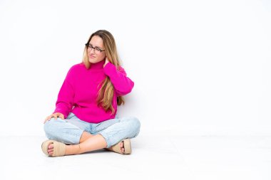 Beyaz arka planda boynu ağrıyan beyaz bir kadın yerde oturuyor.