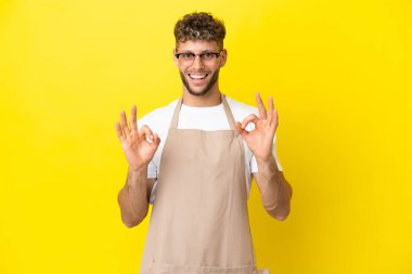 Restaurant waiter blonde man isolated on yellow background showing an ok sign with fingers