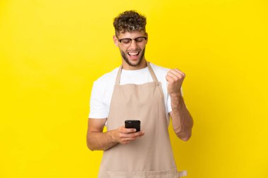 Restaurant waiter blonde man isolated on yellow background with phone in victory position
