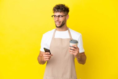 Restaurant waiter blonde man isolated on yellow background holding coffee to take away and a mobile while thinking something