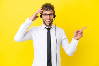 Telemarketer blonde man working with a headset isolated on yellow background surprised and pointing finger to the side