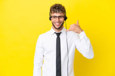 Telemarketer blonde man working with a headset isolated on yellow background making phone gesture. Call me back sign