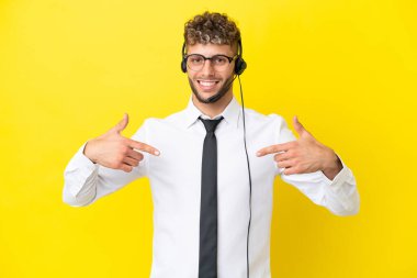 Telemarketer blonde man working with a headset isolated on yellow background proud and self-satisfied