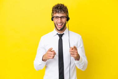 Telemarketer blonde man working with a headset isolated on yellow background pointing to the front and smiling