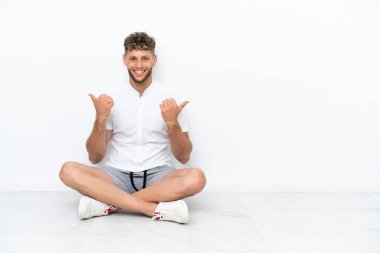 Young blonde man sitting on the floor isolated on white background with thumbs up gesture and smiling