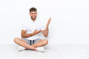 Young blonde man sitting on the floor isolated on white background extending hands to the side for inviting to come