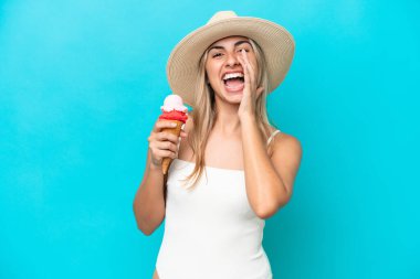 Young caucasian woman in swimsuit with a cornet ice cream isolated on blue background shouting with mouth wide open