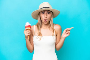 Young caucasian woman in swimsuit with a cornet ice cream isolated on blue background surprised and pointing side