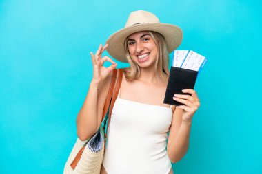 Young caucasian woman in swimsuit holding a passport isolated on blue background showing ok sign with fingers
