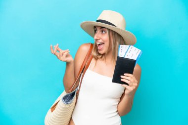 Young caucasian woman in swimsuit holding a passport isolated on blue background with surprise facial expression
