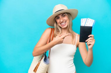 Young caucasian woman in swimsuit holding a passport isolated on blue background and pointing it