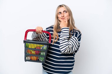 Young caucasian woman holding a shopping basket full of food isolated on white background thinking