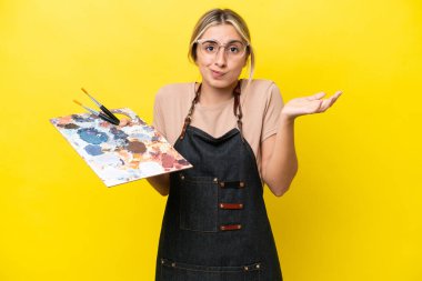 Young artist caucasian woman holding a palette isolated  on yellow background having doubts while raising hands
