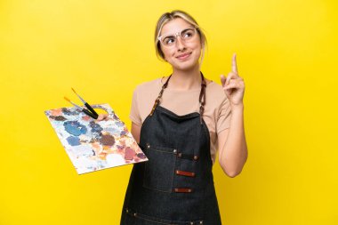 Young artist caucasian woman holding a palette isolated  on yellow background pointing up a great idea