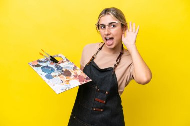 Young artist caucasian woman holding a palette isolated  on yellow background listening to something by putting hand on the ear