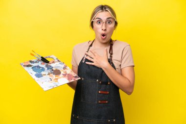 Young artist caucasian woman holding a palette isolated  on yellow background surprised and shocked while looking right