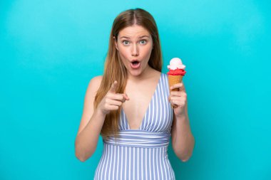 Young caucasian woman in swimsuit eating ice cream isolated on blue background surprised and pointing front